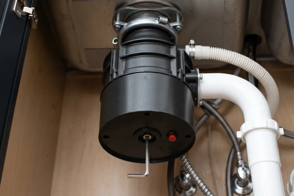 The underside of a garbage disposal with a hex key in the center socket and the red reset button beside it